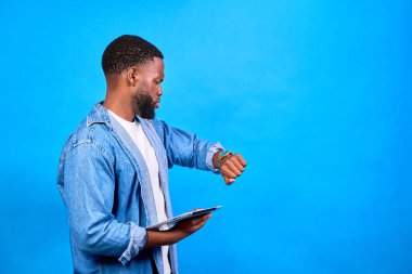 Genç profesyonel elinde bir tablet tutuyor ve saatine bakıyor. Yoğun programını canlı mavi bir arka planla yönetiyor.