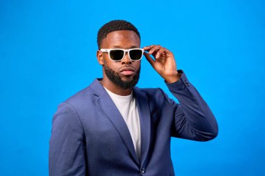 Kendine güvenen iş adamı güneş gözlüğünü ayarlıyor, modern tarzı sergiliyor ve enerjik arkaplana karşı girişimci ruhunu sergiliyor.