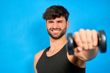 Genç adam ağırlık kaldırırken komik bir ifade takınıyor, parlak mavi bir arka planda izole edilmiş, spor yaparken mizah gösterisi yapıyor.