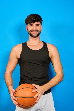 Gülümseyen basketbolcu, canlı, mavi bir arka plana karşı spor kıyafetleriyle güven içinde poz veriyor. Mutluluk ve enerji saçıyor.