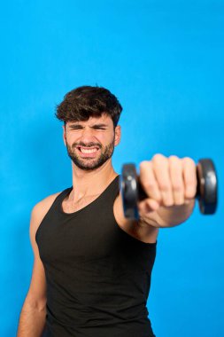 Genç bir adam mavi bir arka planda izole edilmiş, yüzünde komik bir ifadeyle bir dambılı kaldırmakta zorlanıyor.