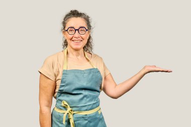 Önlük ve gözlük takan olgun, arkadaş canlısı bir kadın avucunun içinde gri arka planda izole edilmiş bir şey gösteriyor.