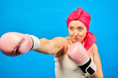 Orta yaşlı, pembe boks eldiveni ve başörtüsü takan bir kadın, kanserle mücadeleyi simgeleyen bir yumruk atıyor.