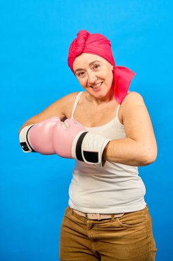 Orta yaşlı, pembe boks eldiveni ve başörtüsü takan bir kadın. Kanserle mücadelesini sembolize ediyor.