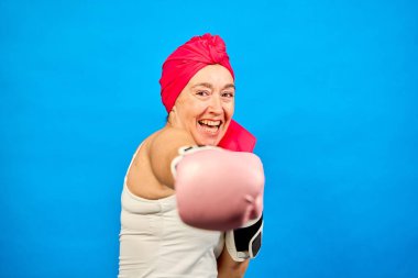 Mavi arka planda gülümseyen orta yaşlı bir kadının pembe boks eldiveni ve başörtüsü taktığı, kanseri yumrukladığı ve dövüştüğü bir fotoğraf.