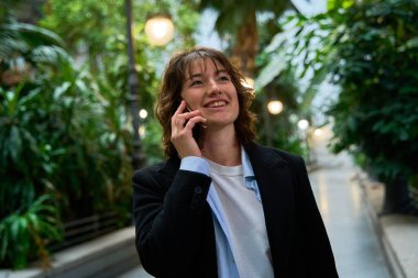 Genç iş kadını akıllı telefonuyla konuşurken gülümsüyor, iş molasında güzel bir kapalı bahçede yürüyor.