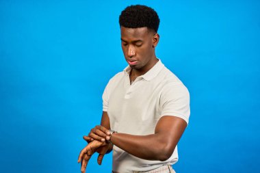 Şık bir genç adam, saati canlı bir mavi arka planla karşılaştırıyor. Dakiklik ve modern yaşam tarzını aktarıyor.