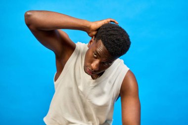 Genç siyahi bir adam saç stilini düzeltirken, temiz, modern bir stüdyo ortamında canlı bir mavi zemine karşı güvenle poz veriyor.
