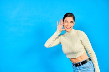 Meraklı genç bir kadın, el ele tutuşuyor, bir konuşma duymaya çalışıyor, canlı mavi bir arkaplana karşı hafif bir sesi dinliyor ya da dinliyor.