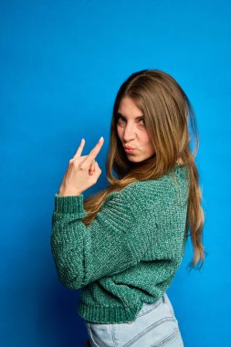 Genç bir kadın parmaklarıyla küçük bir jest yapıyor, izleyiciye bakıyor, yeşil bir kazak ve kot pantolon giyiyor, mavi bir arka planda.