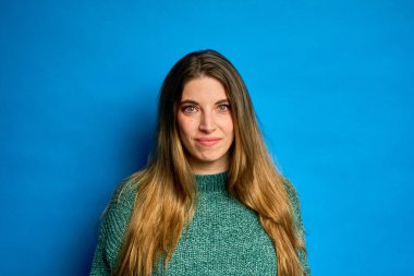 Stüdyo portresi, yeşil kazak giyen, parlak mavi bir arka plana hafifçe gülümseyen uzun saçlı genç bir kadın.