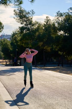 Sportif kadın güneşli bir parkta paten kayıyor, kulaklık takıyor ve spor yaparken eğleniyor.