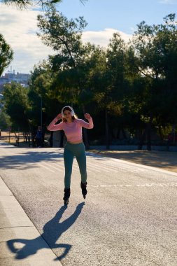 Güneşli bir parkta paten kayan sağlıklı bir kadın, kulaklıkla müzik dinlerken aktif ve sağlıklı bir yaşam tarzını benimsiyor.