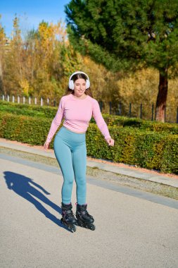 Güneşli bir sonbahar gününde şehir parkında, kablosuz kulaklıklarla patenle gezen ve müzik dinleyen mutlu sporcu kadınlar.
