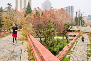 İki adam şehir parkında koşuyor, rengarenk sonbahar yapraklarıyla çevrili olarak antrenmanlarının tadını çıkarıyorlar.