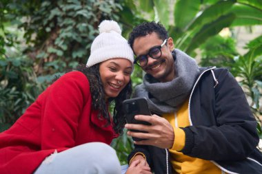 Genç bir çift botanik bahçesinde gülüyor ve cep telefonu kullanıyor. Birlikte mutluluk ve bağlantının tadını çıkarıyorlar.