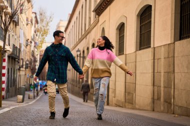 Young multi-ethnic couple holding hands, running and having fun exploring a new city during their european vacation clipart