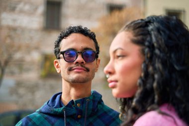 Güneş gözlüklü ve ekose gömlekli genç bir adam kıvırcık saçlı genç bir kadının yanında duruyor. Şehir ortamında gerginlik ve mesafeyi vurguluyor.