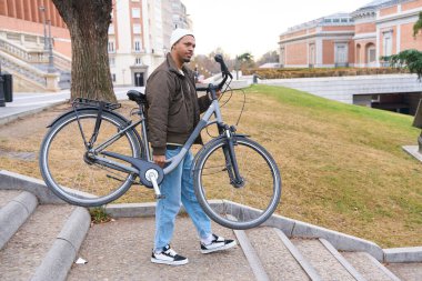 Bisikletini taşıyan genç bir adam şehir parkında merdivenlerden çıkarken, arka planda bir müze ile