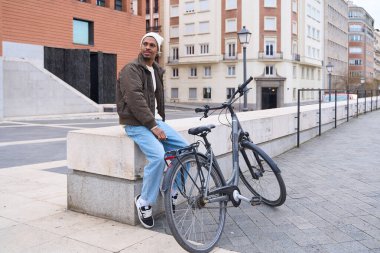 Genç adam şehirde alçak bir duvarda mola veriyor, bisikleti yanında dinleniyor, şehir manzarasının tadını çıkarıyor.