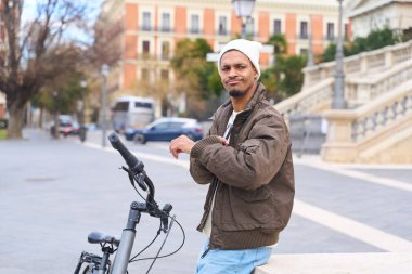 Beyaz bereli ve kahverengi ceketli genç adam, şehir meydanında, elektrikli bisikletiyle alçak bir duvarda oturuyor.