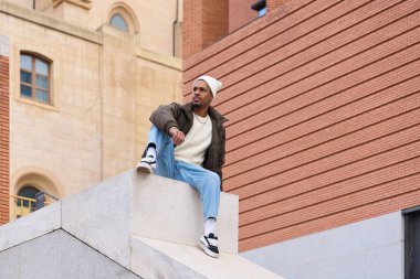 Şık kış kıyafetleri giyen genç adam şehir ortamında alçak bir duvarda oturuyor, çağdaş tarz ve kentsel rahatlama karışımı sergiliyor.