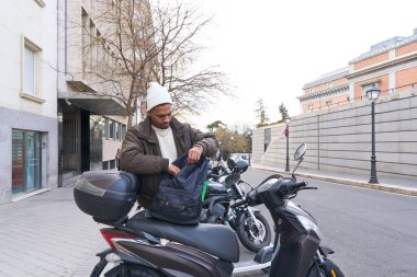 Genç teslimatçı şehirde scooterına yaslanırken termal sırt çantasında bir şey arıyor.