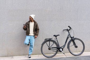 Genç adam, yanında bisikleti olan bir duvara yaslanırken cep telefonu kullanıyor.