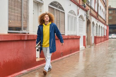 Kıvırcık saçlı genç adam ıslak bir sokakta elinde kaykayla güvenle yürüyor. Kırmızı ve beyaz binalarla kentsel arkaplan