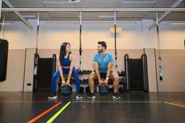 İki fitness meraklısı modern bir spor salonunda kettlebell egzersizi yapıyorlar. Enerjik bir ortamda güç ve dostluk gösteriyorlar.
