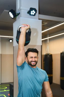 İyi aydınlatılmış bir spor salonunda kettlebell ile egzersiz yapan atletik bir adam kum torbaları ve modern ekipmanlar arasında güç ve zindelik gösterisi yapıyor.