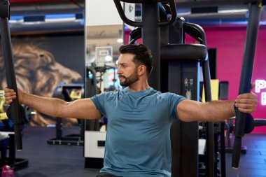 Modern bir spor salonunda göğüs uçuşu makinesi üzerinde çalışan odaklanmış bir adam, dayanıklılık eğitimi ve kendini adamışlığa vurgu yapıyor.