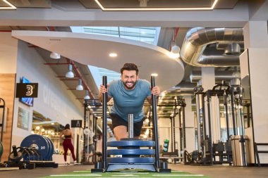 Çağdaş bir spor salonunda ağırlıklı bir kızağı iten enerjik bir adam. Arka planda fitness ekipmanları ve endüstriyel tasarım elemanları