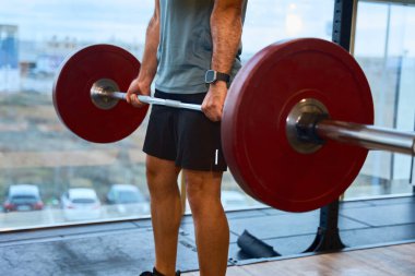Odaklanmış sporcu geniş pencereli bir spor salonunda ağırlık kaldırıyor. Forma ve ağırlık kaldırmaya olan bağlılığını ve gücünü gösteriyor.