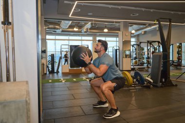 Atletik bir adam modern bir spor salonunda bir tıp topuyla çömeliyor. Güç ve dayanıklılık eğitimine odaklanıyor.