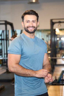 Modern bir spor salonunda güvenle gülümseyen, mavi atletik gömlek giyen, sağlıklı bir yaşam tarzı ve formda olma hevesini sergileyen sakallı, zinde bir adam.
