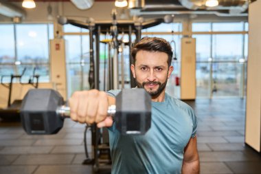 İyi donanımlı bir spor salonunda dambılla egzersiz yapan bir adam. Güç ve kendini spora adamış. Parlak, ferah ortam motivasyonu artırır