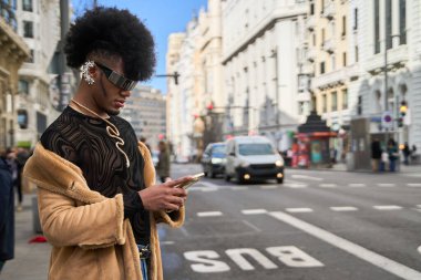 Şehir caddesinde otobüs durağında beklerken akıllı telefon kullanan afro saç stili olan şık bir insan.