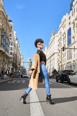 Şehir caddesinde bej ceketli, yüksek topuklu ve kot pantolonlu afro saçlı şık bir adam.