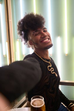Afro saçlı, çift cinsiyetli gülümseyen genç adam, neon ışıklarla dolu modern bir barda birayla selfie çekiyor.