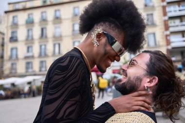 İki çift cinsiyetli olmayan insan bir şehir meydanında kucaklaşıp gülümsüyor, aşklarını ve kimliklerini kutluyorlar.