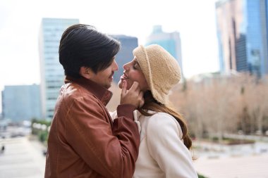 Erkek kadının yüzünü nazikçe okşar, arka planda şehir manzarasıyla samimi bir anı paylaşır.