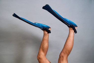 Mavi dalış paletleri giyen kıllı erkek bacakları gri bir arka plana doğru yükselmiş ve sualtı keşiflerini çağrıştırıyor.