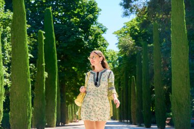 İspanya, Seville 'deki Maria Luisa Parkı' nda yürüyen genç bir kadın selvi ağaçlarıyla çevrili güneşli güzel günün tadını çıkarıyor.