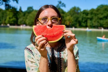Yüzünün önünde karpuz dilimi tutan neşeli genç kadın gölün kenarında güneşli bir günün tadını çıkarıyor.