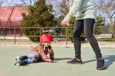 Parkta güneşli bir günde annesiyle paten kayarken koruyucu kask takmış gülümseyen küçük bir kız.