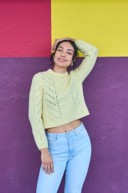 Açık sarı kazaklı ve açık mavi kotlu gülümseyen genç bir kadın elini renkli bir duvarın üzerine koyup poz veriyor.