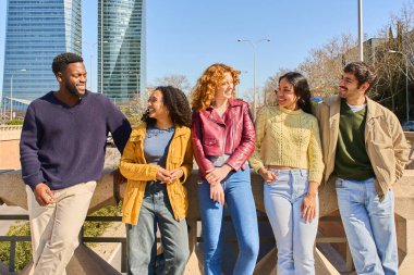 Şehirdeki bir köprüde sohbet eden beş genç öğrenci, arka planda modern gökdelenler var.