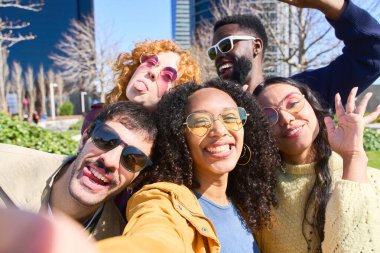 Bir grup neşeli, çok ırklı arkadaş dışarıda selfie çekmek için poz veriyor, aptal suratlar yapıyor ve parkta güneşli bir günün tadını çıkarıyorlar.