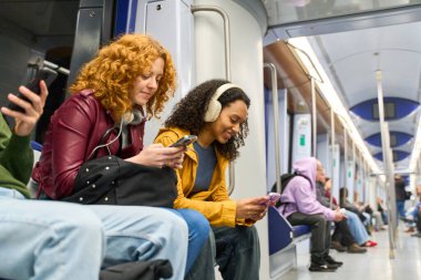 İki genç kadın metroda oturuyorlar, akıllı telefonlarını kullanıyorlar ve günlük yolculukları sırasında kulaklıkla müzik dinliyorlar.